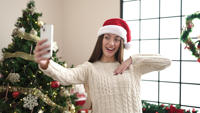 Young beautiful hispanic woman standing by christmas tree make selfie by camera at home
