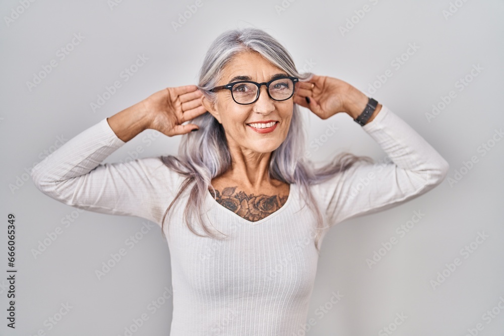 Wall mural middle age woman with grey hair standing over white background smiling pulling ears with fingers, fu