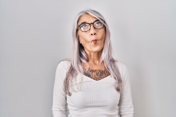 Middle age woman with grey hair standing over white background making fish face with lips, crazy...