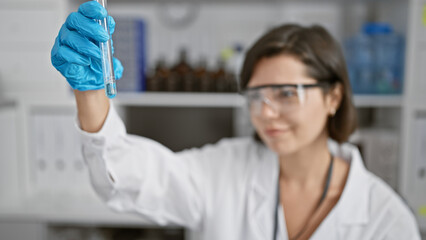 Confident & beautiful young hispanic woman scientist, smiling while holding a test tube, hard at work in a bustling laboratory setting, basking in the glow of her radiant experiments.