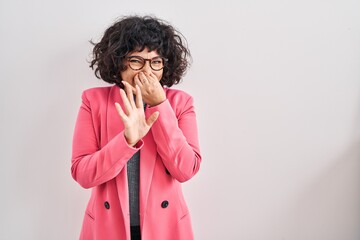 Hispanic woman with curly hair standing over isolated background smelling something stinky and...