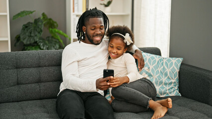 African american father and daughter using smartphone sitting on sofa smiling at home