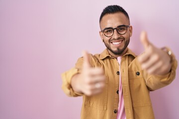 Naklejka premium Young hispanic man standing over pink background approving doing positive gesture with hand, thumbs up smiling and happy for success. winner gesture.