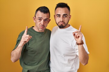 Homosexual couple standing over yellow background pointing up looking sad and upset, indicating...