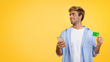 Young European man with credit card and cellphone on yellow studio backdrop