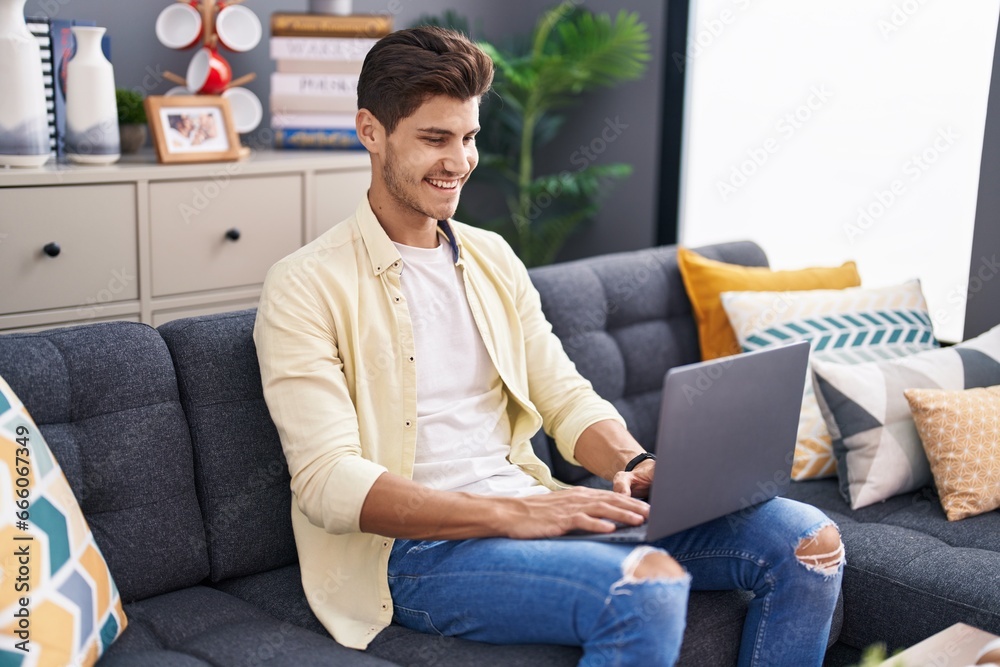 Wall mural Young hispanic man using laptop sitting on sofa at home