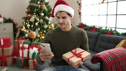 Obraz na płótnie Canvas Young hispanic man using smartphone sitting on sofa by christmas tree at home