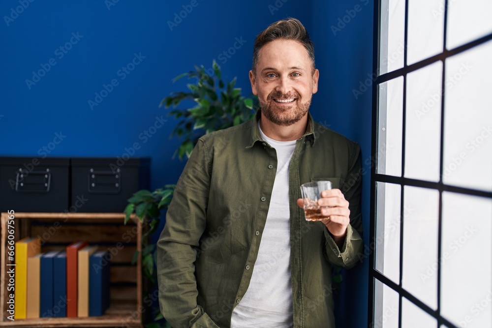 Sticker young caucasian man drinking glass of whisky standing at home