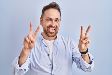 Middle age caucasian man standing over blue background smiling with tongue out showing fingers of both hands doing victory sign. number two.