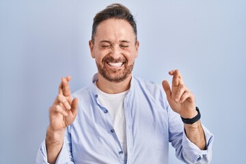 Middle age caucasian man standing over blue background gesturing finger crossed smiling with hope and eyes closed. luck and superstitious concept.