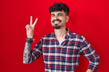 Naklejka premium Young hispanic man with beard standing over red background smiling looking to the camera showing fingers doing victory sign. number two.