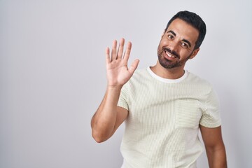 Hispanic man with beard standing over isolated background waiving saying hello happy and smiling, friendly welcome gesture