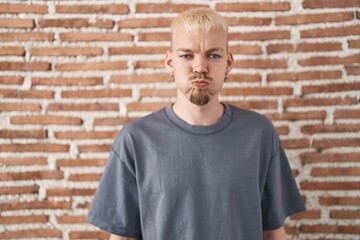 Young caucasian man standing over bricks wall puffing cheeks with funny face. mouth inflated with...