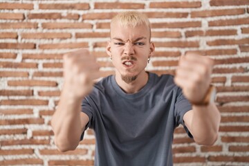 Young caucasian man standing over bricks wall angry and mad raising fists frustrated and furious while shouting with anger. rage and aggressive concept.