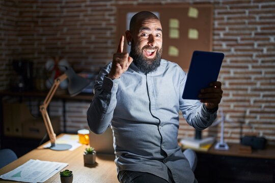 Young hispanic man with beard and tattoos working at the office at night pointing finger up with successful idea. exited and happy. number one.