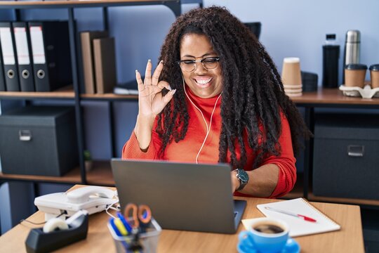 Plus size hispanic woman working at the office with headphones doing ok sign with fingers, smiling friendly gesturing excellent symbol