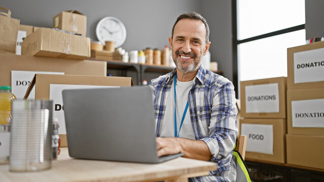 Smiling middle-aged man with grey hair confidently volunteers at a community charity center, exuding unity and altruism while skillfully operating a laptop.