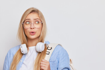 Blond girl in glasses and with earphones on neck posing on white background looking surprised, busy day concept, copy space