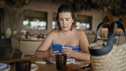 Young hispanic woman using smartphone sitting on the table at the restaurant