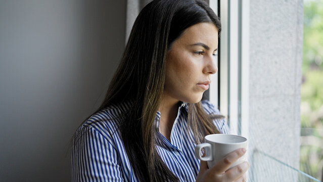 Young beautiful hispanic woman standing looking through the window with sad expression at home