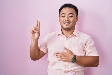 Chinese young man standing over pink background smiling swearing with hand on chest and fingers up,...