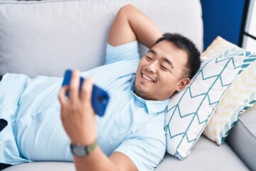 Young chinese man using smartphone lying on sofa at home