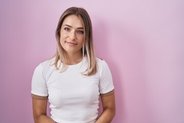 Blonde caucasian woman standing over pink background with hands together and crossed fingers smiling relaxed and cheerful. success and optimistic