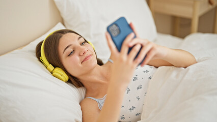 Obraz na płótnie Canvas Young beautiful girl using smartphone and headphones lying on bed at bedroom