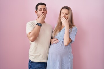 Young couple expecting a baby standing over pink background bored yawning tired covering mouth with...