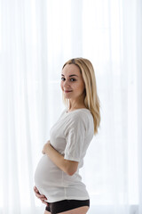 A young blonde pregnant woman stands by the window and strokes her belly in the rays of the morning sun. tender photo session of the expectant mother. pregnant woman in a short top and shorts