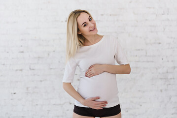 A pregnant woman in a white T-shirt and short shorts stands near a large panoramic window with a light tulle curtain and gently touches her belly. Future mother expecting a baby