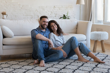 Happy attractive young adult husband and wife resting on heating clean carpeted floor, sitting close, looking at camera, smiling, laughing. Married couple of new homeowners in love home portrait - obrazy, fototapety, plakaty