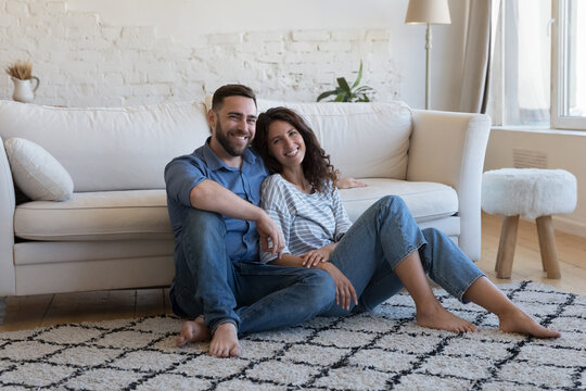 Happy attractive young adult husband and wife resting on heating clean carpeted floor, sitting close, looking at camera, smiling, laughing. Married couple of new homeowners in love home portrait