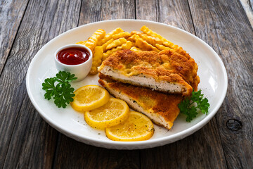 Crispy breaded seared chicken cutlet with French fries, lemon and fresh vegetables on wooden table
