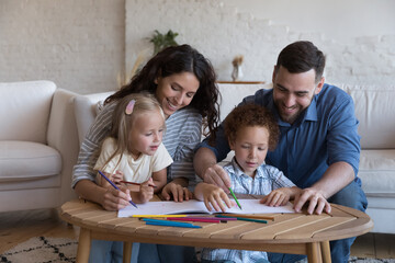 Cheerful mom and dad teaching two cute little kids to draw in colored pencils, having fun,...