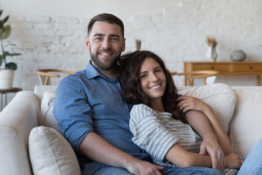 Happy attractive millennial married couple home portrait. Handsome boyfriend and beautiful girlfriend, husband and wife hugging on cozy sofa at new home, looking at camera, smiling