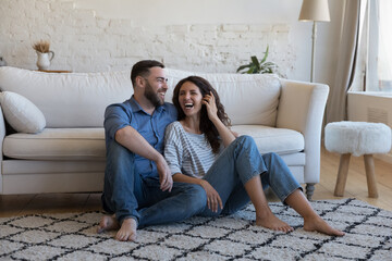 Cheerful carefree married couple in love resting on warm carpeted floor in cozy living room,...