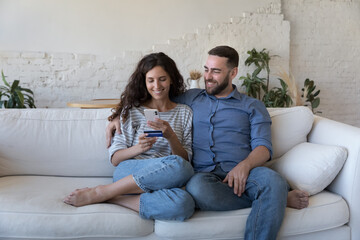 Happy cheerful millennial couple sharing mobile phone and credit card, shopping online, using...