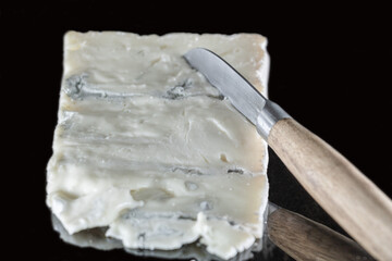 Fromage italien Gorgonzola sur un fond noir avec effet miroir et un couteau à fromage avec un manche en bois posé dessus, différentes couches de bleu et crème apparentes  - obrazy, fototapety, plakaty