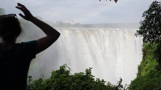 Slow motion footage of The great Victoria Falls waterfall in Zimbabwe at the border with Zambia. New 7 Natural Wonders. Mosi-oa-Tunya on the river Zambezi with a rainbow and silhouette of a woman.