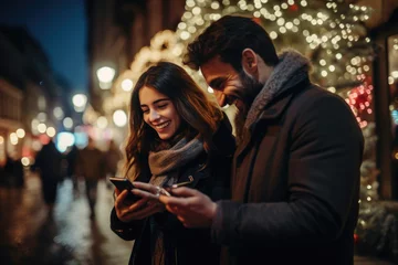 Fotobehang Amidst the vibrant city lights during Christmas and New Year, a couple radiates happiness while holding a mobile phone, their smiles lighting up the night © Konstiantyn Zapylaie