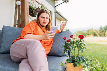 Young redhead woman relaxing with phone in the yard of the house in summer. Negative people...