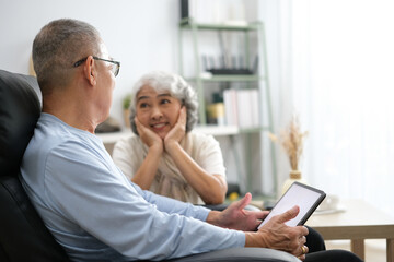 senior couple sitting on sofa using tablet while video call online with family in living room at home