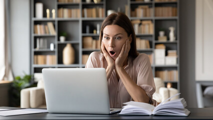 Amazed woman seated at desk in cozy modern living room gawp at laptop screen feels shocked about...