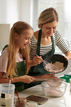 Cheerful mother and daughter sifting seasoned flour in big bowl when making gingerbread dough