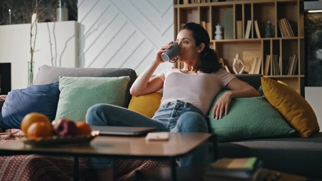 Relaxed woman freelancer drinking tea sitting cozy couch living room alone. 