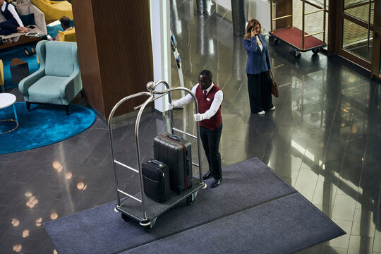 High angle view of African American doorman carrying the trolley with luggage in lobby of the hotel