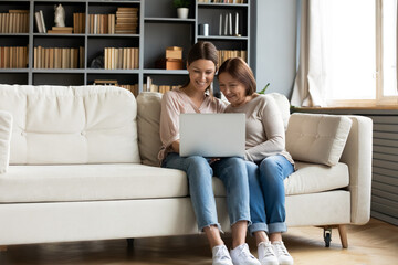 Women on-line buyers enjoy internet shopping seated together on couch at home, grown up daughter hold on lap pc explain to mother new apps, ecommerce users, personal development or having fun concept