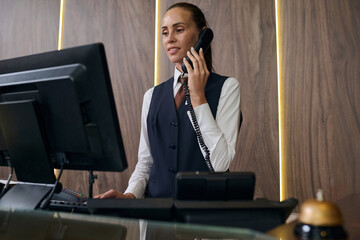 Manager in uniform standing behind the counter with computer and registrating guest by the phone