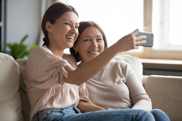 Beautiful adult daughter and middle-aged mum sitting on sofa making photo having fun at home,...